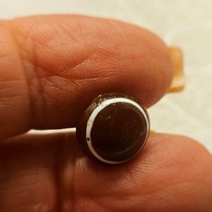 Puede incluir: Una pequeña cuenta redonda, de color marrón oscuro, con una franja blanca alrededor del borde. La cuenta se sostiene entre dos dedos, mostrando su tamaño y diseño. La superficie lisa y los colores contrastantes crean una estética simple pero elegante.