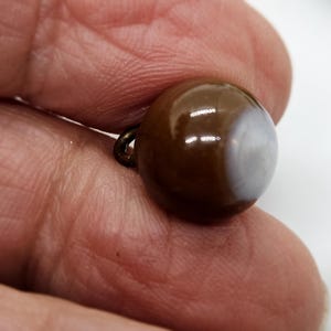 Puede incluir: Un botón redondo marrón con una sección blanca veteada. El botón tiene un pequeño lazo de metal adjunto en la parte posterior. El botón es liso y brillante.