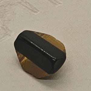 May include: A polished gemstone with a black, rectangular center set on a golden-brown base. The stone has a smooth, reflective surface. The overall shape is irregular, with a natural, organic appearance. This could be a pendant or a bead.