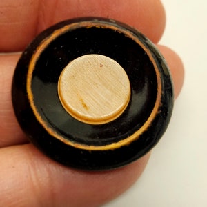 May include: A black and gold round button with a cream-colored center. The button has a raised edge and a smooth surface.