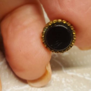 Puede incluir: Un pequeño botón redondo con un centro negro y un borde dorado texturizado. El botón se sostiene entre los dedos, mostrando su tamaño y diseño. La superficie del botón refleja la luz, resaltando su acabado pulido.