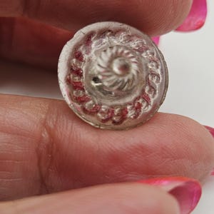 Puede incluir: Un botón redondo y transparente con un diseño en espiral en el centro y un borde de detalles rojos. El botón se sostiene entre los dedos, mostrando sus intrincados detalles y su estética vintage. El botón probablemente está hecho de vidrio o un material transparente similar.