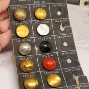 Puede incluir: Una tarjeta de botones vintage con una variedad de botones en diferentes colores y acabados. Los botones incluyen plata, oro, negro, rojo y níquel. La tarjeta está etiquetada con la talla 30 y varios códigos.