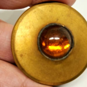 May include: A vintage brass button with a central amber-colored glass cabochon. The button has a circular design with a raised rim and a slightly tarnished finish, suggesting age and use. The cabochon is round and faceted.