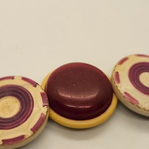 May include: Three round game pieces, one is a solid dark red color, the other two are white with concentric circles of purple.