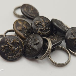 Peut inclure: Un ensemble de boutons anciens de couleur laiton avec un emblème de scout et l'inscription "BE PREPARED" en relief. Chaque bouton est attaché à un petit anneau métallique argenté. Les boutons sont dispersés sur une surface blanche.
