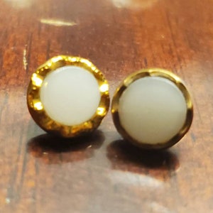 Puede incluir: Un par de pendientes de botón dorados con un centro blanco y circular. Los pendientes tienen un diseño de borde festoneado. Los pendientes se muestran sobre una superficie de madera.
