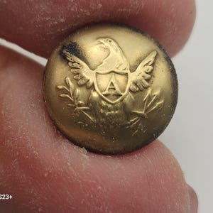 May include: A round, antique brass button with an embossed eagle design. The eagle is centered above a shield with the letter "A" inside. The button has a slightly worn appearance, suggesting age and use. The button is held between fingers.