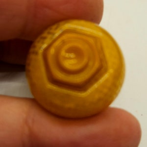 May include: A close-up of a gold-colored plastic hexagonal button with a raised, concentric circle design.