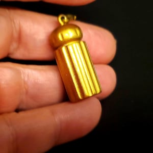 Puede incluir: Un colgante dorado con un cuerpo cilíndrico y una parte superior redondeada. El colgante tiene estrías verticales a lo largo de su cuerpo y un pequeño lazo en la parte superior para una cadena. El fondo es negro.