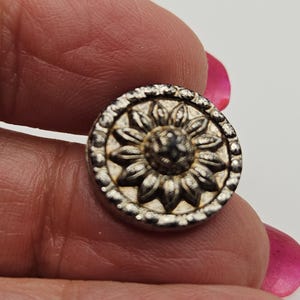 May include: A small, round, silver-toned button with a raised sunflower design in the center. The button has a detailed, textured border. The button is held between two fingers.