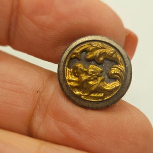 May include: A small, round button with a gold-colored, ornate design set within a silver-toned border. The gold design features a detailed, possibly floral or mythical, motif. The button is likely made of metal and has a vintage or antique appearance.