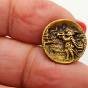 May include: A round, antique brass button with a detailed relief of a figure playing a horn, a castle, and a boat. The button has a vintage aesthetic and is held in a hand.