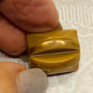 May include: A square, mustard-colored button with a carved, curved design. The button is held between fingers, showcasing its glossy finish and detailed texture. This vintage button is a craft supply.