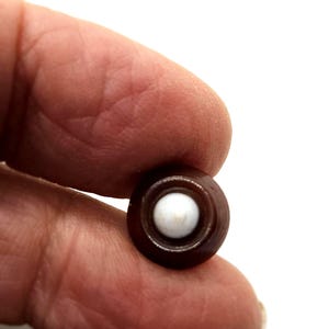 Puede incluir: Un botón pequeño, redondo, de color marrón oscuro con un centro blanco y circular. El botón se sostiene entre los dedos sobre un fondo blanco. El botón es probablemente para coser o manualidades.