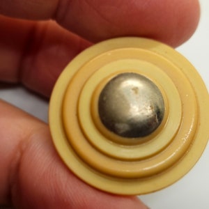 May include: A vintage button with a layered design in shades of yellow and tan, featuring a silver-colored, rounded center. The button has a circular shape with concentric rings, creating a textured appearance. This button is a unique craft supply.