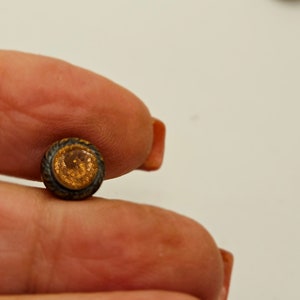 Puede incluir: Primer plano de un botón pequeño, redondo, de color dorado con un borde negro. El botón tiene una superficie texturizada y parece estar hecho de metal.