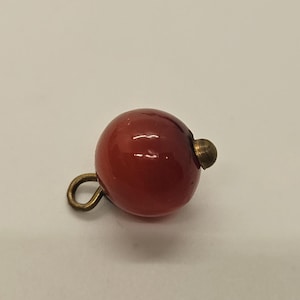 May include: A round, glossy, red bead with a brass-colored loop and a small brass-colored stud. The bead is a deep, rich red color and appears to be made of a smooth, polished material.