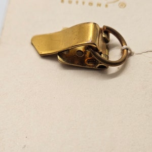 Könnte beinhalten: Ein goldfarbener Metallverschluss mit einem Ring. Der Verschluss ist zum Befestigen an einem Kleidungsstück gedacht.