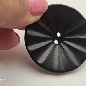Könnte beinhalten: Ein großer, schwarzer Knopf mit radialem Design und zwei Löchern. Der Knopf wird von einer Person mit rosa Nagellack gehalten. Das Design des Knopfes weist eine Reihe von gekrümmten Linien auf, die vom Zentrum ausgehen und eine strukturierte Oberfläche bilden.