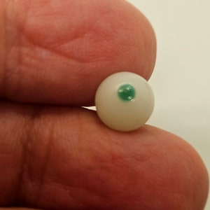 Puede incluir: Un ojo pequeño, redondo y blanco con iris verde. El ojo es una réplica en miniatura, probablemente para manualidades o muñecas. El ojo se sostiene entre dos dedos, mostrando su tamaño y detalle.