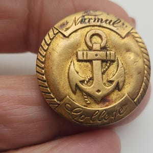 May include: A vintage gold-colored button with an anchor design. The button has the words "Normal" and "College" inscribed on it. The button is round with a rope-like border.