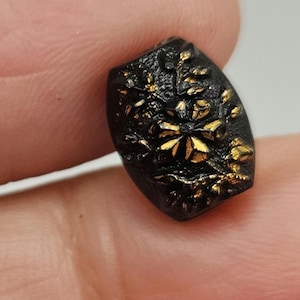 May include: A small, black, oval-shaped button with a floral design in gold. The button has a textured surface and is held between two fingers. The gold floral design is centered on the button and is surrounded by smaller gold accents.