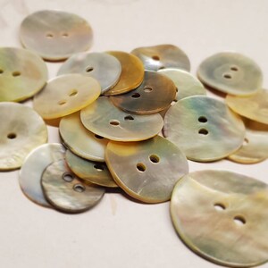May include: A selection of iridescent, circular buttons, each with two holes. The buttons display a natural, pearlescent lustre, with hues from pale yellow to brown and green. Suitable for sewing and craft activities.