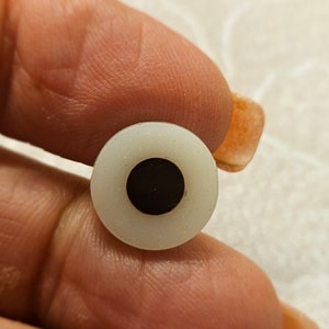 Puede incluir: Un ojo de muñeca de plástico blanco con una pupila negra. El ojo tiene un diámetro de aproximadamente 2,5 cm.