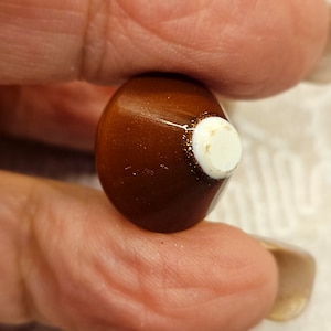 Può includere: Primo piano di una perla marrone a forma di goccia con un accento circolare bianco. La perla ha una finitura lucida ed è tenuta tra due dita. Un anello di glitter circonda l'accento bianco.