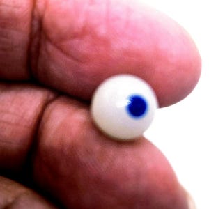 Puede incluir: Un pequeño ojo de muñeca redondo y blanco con un iris azul vibrante. El ojo se sostiene entre los dedos, mostrando su superficie lisa y brillante. El fondo es de un blanco brillante.