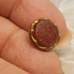 Puede incluir: Una pequeña piedra preciosa redonda, de color marrón rojizo, engastada en un bisel dorado. La piedra preciosa tiene una forma ligeramente irregular y una superficie texturizada. El bisel dorado tiene un diseño ondulado y orgánico. El artículo se sostiene en una mano.