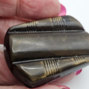 May include: A dark brown, rectangular vintage brooch with a curved top and bottom. The brooch has a smooth, polished surface with etched linear details on the top and bottom edges. The brooch is held in a hand against a white background.