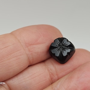 May include: A small, black, carved gemstone with a floral design. The gemstone is square-shaped with rounded corners and features a starburst pattern in the center. The stone is held in a hand.