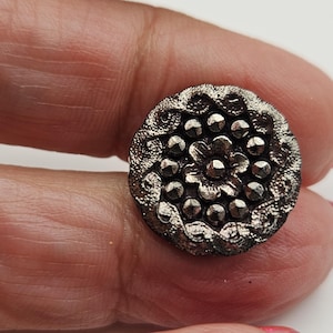 Può includere: Un bottone d'argento antico e dettagliato con un motivo floreale. Il bottone presenta un fiore centrale circondato da punti più piccoli in rilievo e un bordo ornato e strutturato. Il bottone è rotondo e sembra essere di metallo.