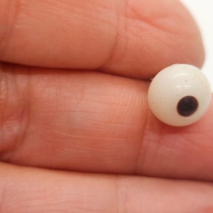 Puede incluir: Una pequeña cuenta esférica blanquecina con un punto circular marrón oscuro. La cuenta se sostiene entre los dedos sobre un fondo neutro. La cuenta es probablemente un componente para joyería o manualidades.