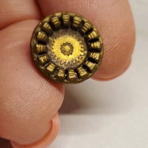 May include: A small, round button with a detailed design. The button has a gold center with a floral pattern and a bronze-colored outer ring with a ridged texture. The button is held between fingers.