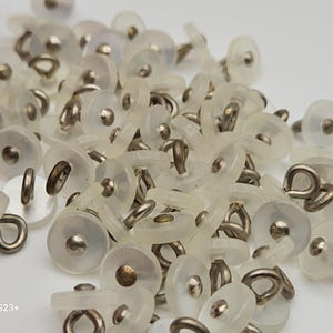 May include: A pile of clear, round buttons with metal shanks. The buttons have a slightly translucent appearance, with a small, silver-colored center. The metal shanks are a silver color and are attached to the back of the buttons.
