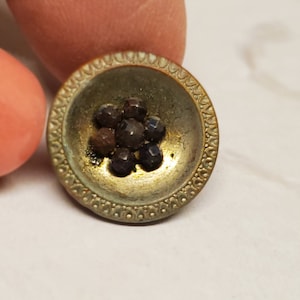 Puede incluir: Un botón vintage con una base de metal dorado y un borde elevado y adornado. El centro del botón presenta un grupo de piedras de vidrio marrón oscuro y negro.