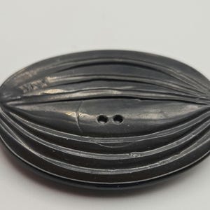 Peut inclure: Un bouton ovale gris foncé avec une surface texturée et deux petits trous. Le bouton présente une série de lignes courbes parallèles qui créent un effet côtelé. Le bouton est probablement en plastique ou dans un matériau similaire.
