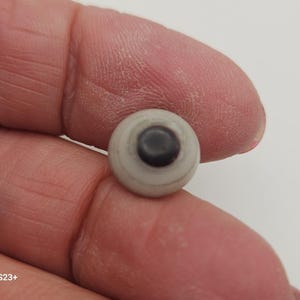 Puede incluir: Un pequeño ojo redondo blanquecino con una pupila negra. El ojo se sostiene entre los dedos sobre un fondo blanco. El ojo es un suministro artesanal o un componente para muñecas u otros artículos hechos a mano.