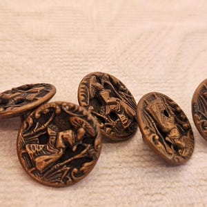 May include: A collection of antique bronze-colored buttons. Each button features a detailed relief design of a figure in a landscape. The buttons are oval-shaped and have a slightly aged appearance, suggesting they are vintage.
