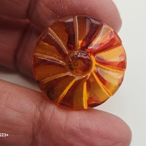 Può includere: Un bottone rotondo color ambra con un design radiale. Il bottone ha un cerchio centrale in rilievo ed è tenuto tra le dita. Il design del bottone presenta segmenti ambra chiari e scuri alternati.