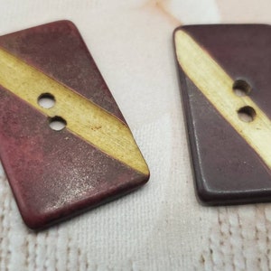 May include: Two rectangular buttons with a diagonal yellow stripe across a maroon background. Each button has two holes for sewing. The buttons are likely made of a hard, durable material.
