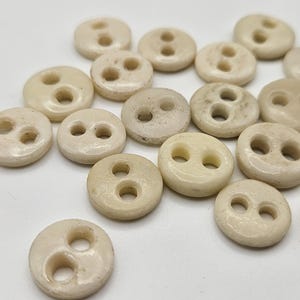 May include: A collection of off-white, round buttons with two holes each. The buttons vary slightly in size and have a smooth, slightly textured surface. They are scattered on a white background, suggesting a craft or sewing project.
