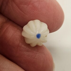 May include: A small, white, flower-shaped bead with a blue center. The bead has a ribbed design and is held between fingers. The bead is likely used for jewelry making or other crafts.