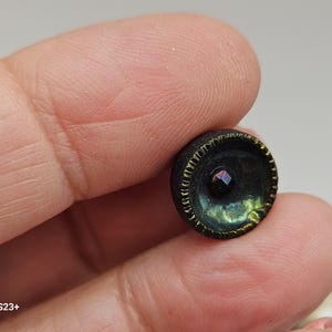 May include: A small, dark button with a textured rim and a central iridescent gem. The button's color shifts from dark green to blue and gold, with a small, faceted gem at its center. The button is held between fingers.