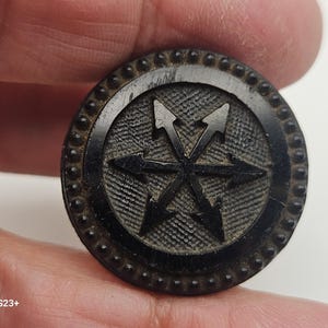 Puede incluir: Un botón negro con un diseño de estrella de seis puntas con flechas que apuntan hacia afuera. El botón tiene una superficie texturizada y un borde con cuentas. El botón se sostiene en una mano.