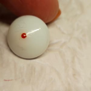 Puede incluir: Canica de vidrio blanco con un pequeño punto rojo en el centro.