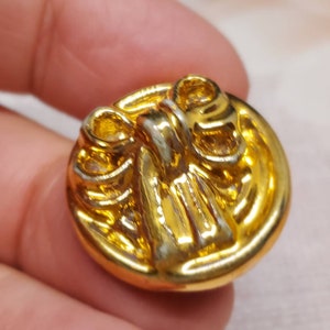 May include: A gold-colored button with a raised bow design. The button is round and has a shiny finish. The bow is in the center of the button and has detailed ridges.